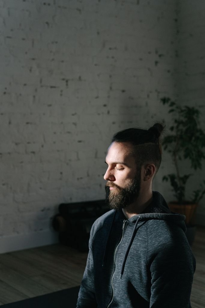 A man with closed eyes and facial hair meditates indoors against a brick wall.