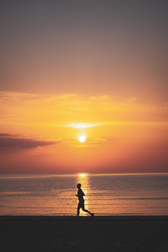 A person jogging on the beach with a stunning sunset, creating a serene silhouette effect.